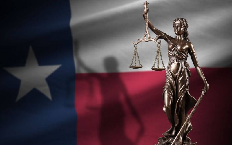 A bronze statue of Lady Justice holding scales set against the Texas state flag background.