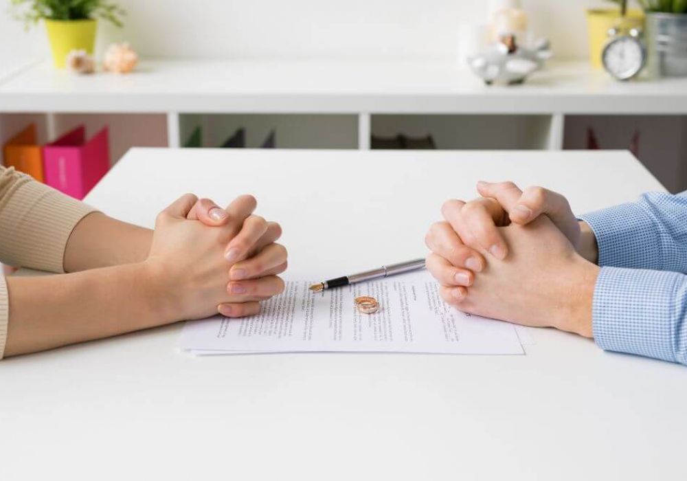 Two people are holding hands across a table with a marriage dissolution document between them, suggesting a couple going through a divorce.