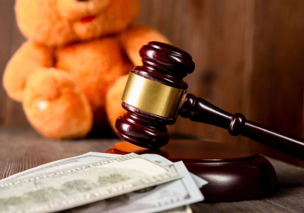 A wooden gavel rests on its stand next to cash, with a plush teddy bear blurred in the background, suggesting legal implications involving children or family.