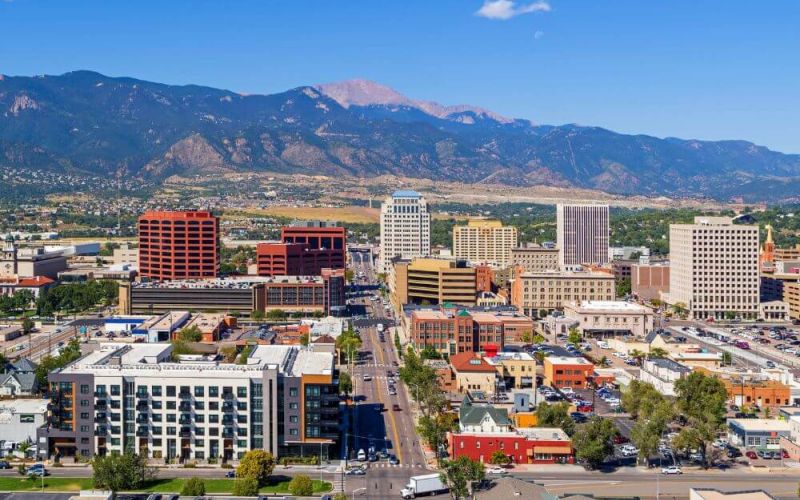 A cityscape showcases a variety of buildings under clear skies, with a backdrop of large mountains and the hint of a daytime moon.