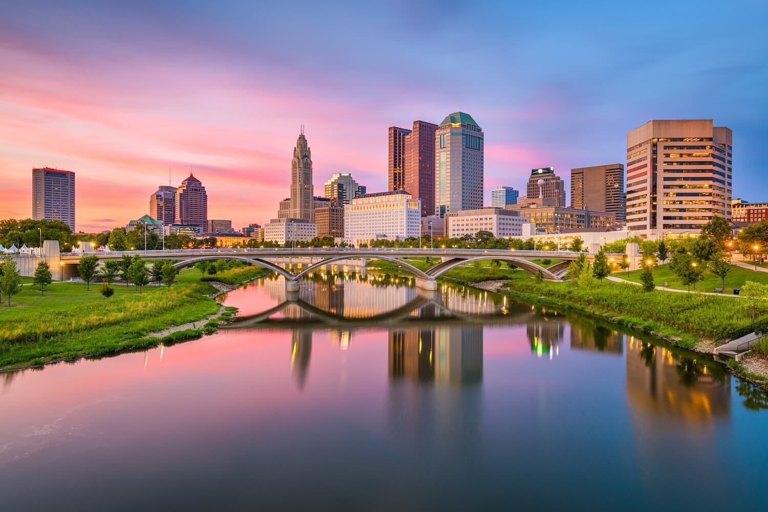 Columbus, Ohio, USA skyline at twilight reflects on a calm river, with an arched bridge in the foreground and colorful clouds above.