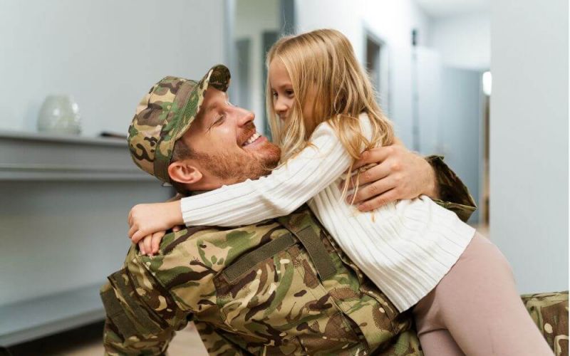 A smiling man in camouflage military attire embraces a joyful young girl in an indoor setting. Their affectionate interaction conveys a warm emotional connection.
