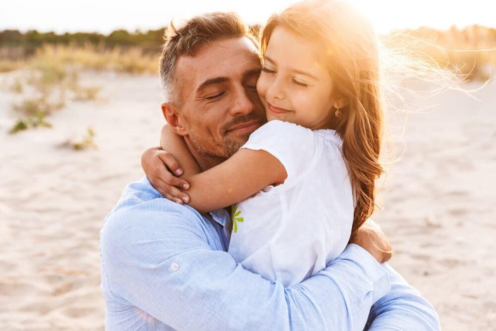 A man embraces a young girl affectionately on a sandy beach at sunset. Both exhibit a content and peaceful demeanor.