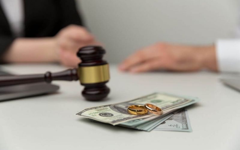 A gavel rests beside two gold wedding rings and a stack of cash on a table, implying a legal issue involving marriage finances, with a partial view of a person's hands in the background.