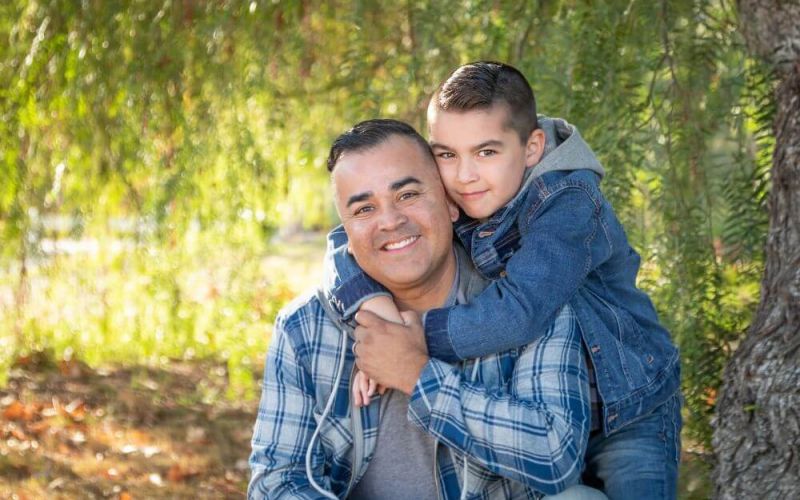 A man and a young boy in denim outfits embrace, smiling in a sunlit, leafy park.