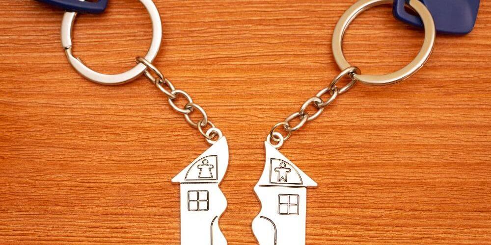 Two keychains with split house-shaped pendants are connected by a chain, resting on a wooden surface, symbolizing joint ownership or separation.