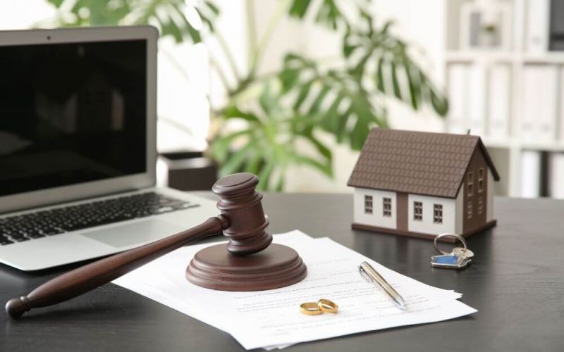 A wooden gavel rests on a document beside a laptop, house model, and keys, implying legal proceedings, possibly real estate, on an office desk.