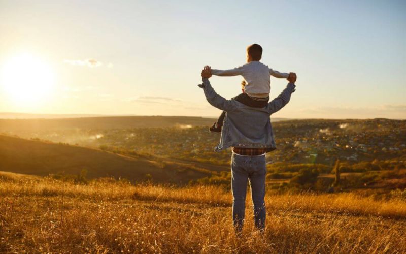 An adult with a child on their shoulders stands in a field at sunset, overlooking a sunlit valley.