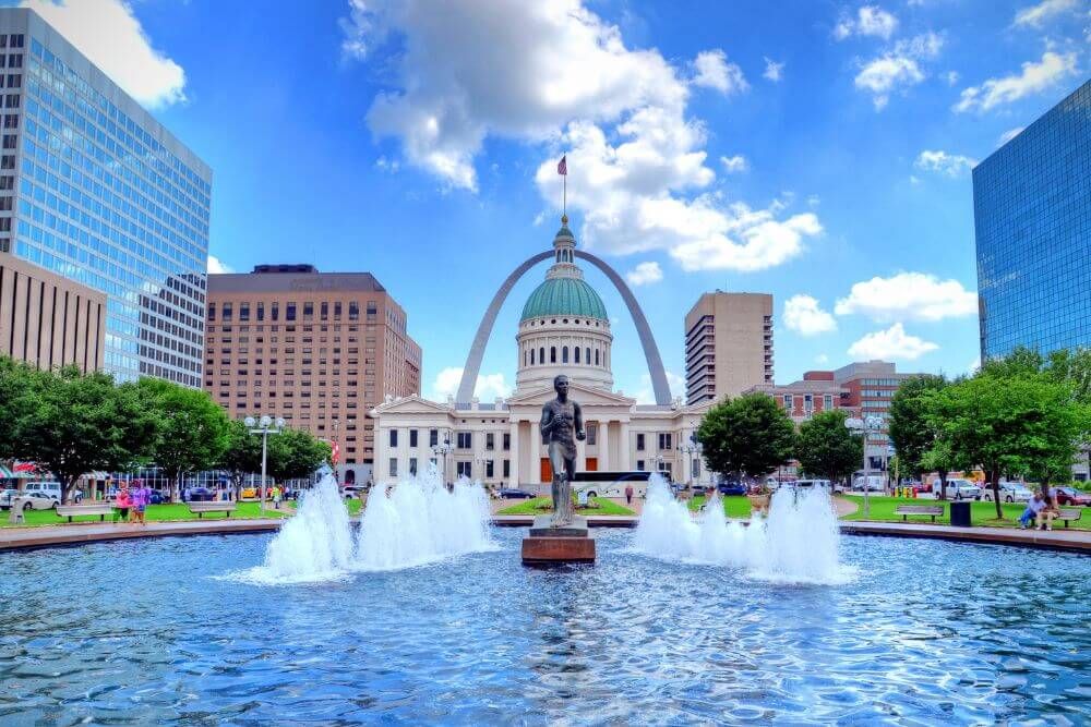 Water fountains gush in a park with people, framed by a domed building and the arching Gateway Arch in a vibrant urban landscape.
