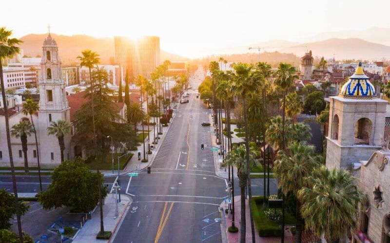 A sunset bathes a cityscape, highlighting palm trees and historical architecture along a wide street, inviting tranquility in a seemingly Californian atmosphere. There is no text in the image.