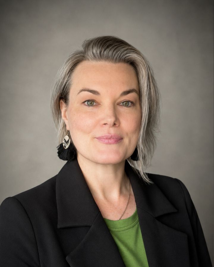 Professional headshot of a smiling woman with short gray hair, wearing a black blazer and green top against a neutral backdrop.
