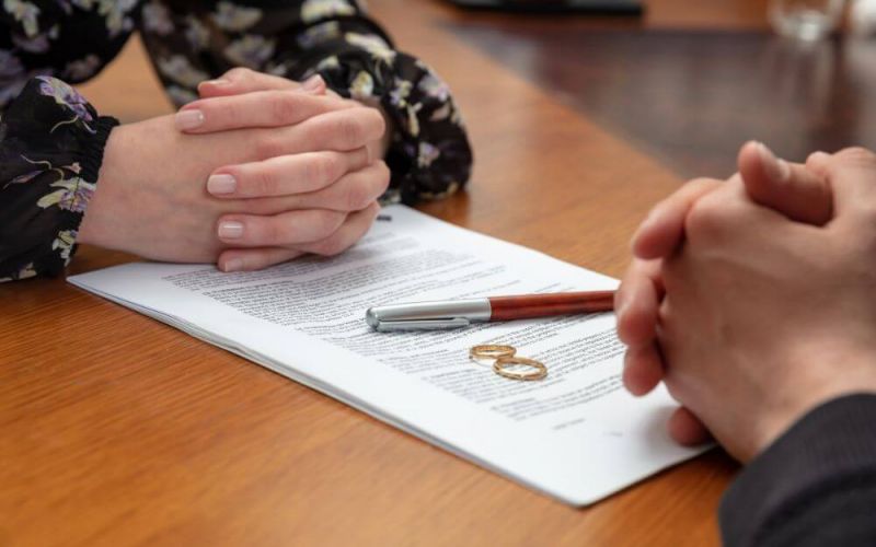 Two hands clasped together on top of a document, with wedding rings and a pen placed beside it, indicating a setting of a formal agreement or contract, likely a marriage.