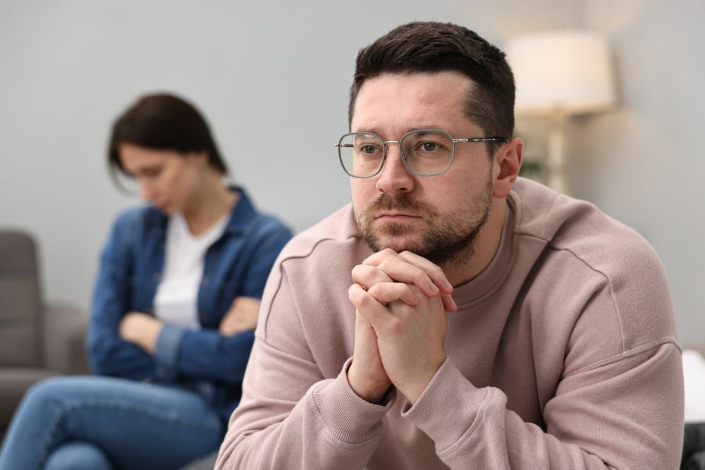 A man with glasses looks contemplative, hands clasped, foreground; a woman sits behind him, out of focus, appearing upset. Context suggests a domestic setting with tension.