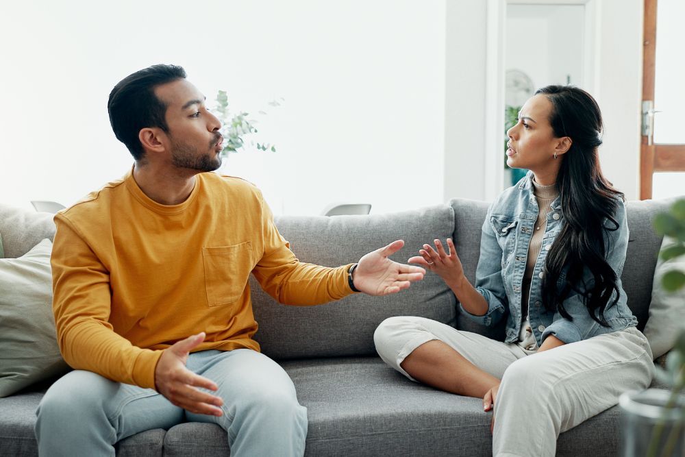 Two people are conversing on a grey couch, gesturing expressively, in a bright living room setting.