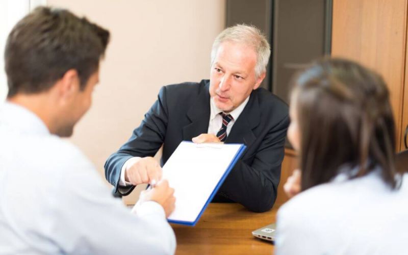 Three individuals in a business meeting, one presenting a document to sign, in an office setting.