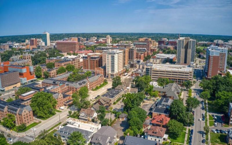 Aerial view of a densely built urban area with a mix of modern high-rises, traditional buildings, tree-lined streets, and a clear blue sky.