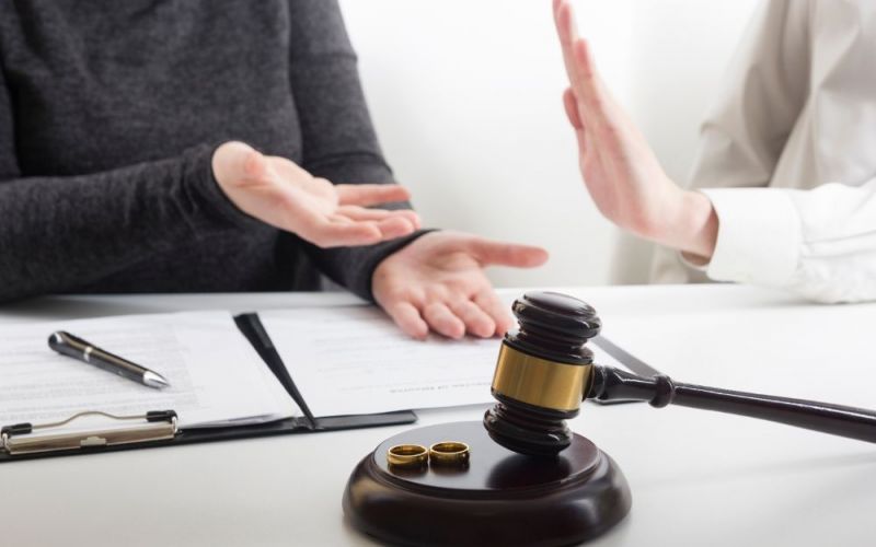 Two people gesticulate during a discussion, with a gavel and wedding rings on the table, suggesting a divorce proceeding context.