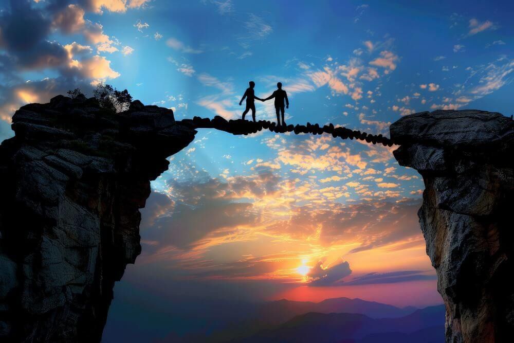 Two silhouetted figures meet halfway on a rope bridge spanning a vast chasm, amidst a dramatic sunset sky with clouds and mountains in the background.