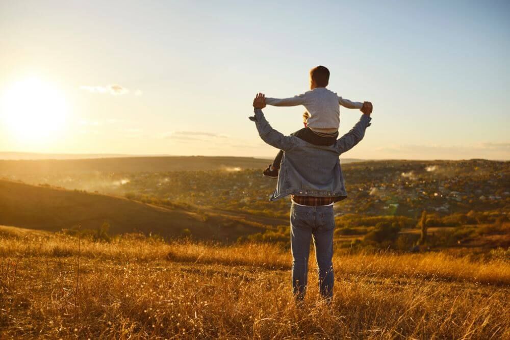 An adult with a child on their shoulders stands in a field at sunset, overlooking a sunlit valley.