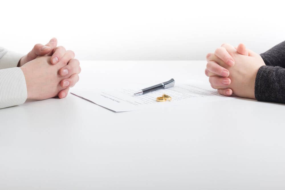 Two people's hands clasped together on a table, with a document, pen, and two wedding rings between them, suggesting a context of either marriage or divorce proceedings.