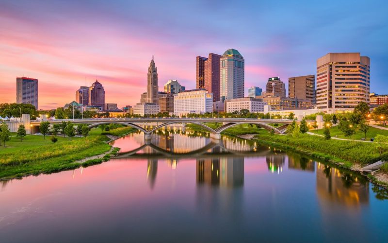 Columbus, Ohio, USA skyline at twilight reflects on a calm river, with an arched bridge in the foreground and colorful clouds above.