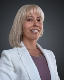 Smiling woman with shoulder-length blonde hair wearing a white blazer and mauve top, posed against a dark gray gradient background.
