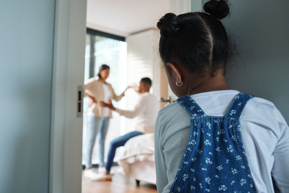 A young girl with double buns looks through a doorway where two adults, blurry in the background, seem to be having a playful moment in a bright room.