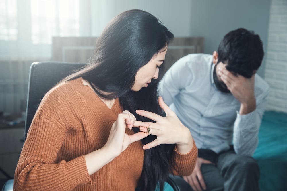 A woman removes a ring from her finger, possibly symbolizing a breakup, while a man covers his face, conveying distress. The setting appears to be an indoor domestic space.