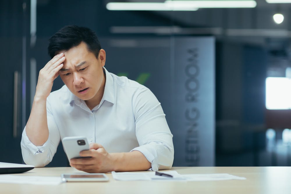 A person appears concerned while looking at a smartphone, hand on forehead, in a modern office with a sign saying "MEETING ROOM." There is no additional text.