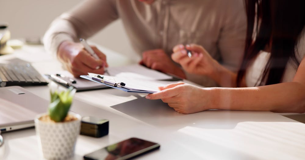 Two individuals are reviewing and signing documents on a clipboard in an office setting with laptops, a smartphone, and desk accessories.