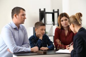 A family attentively listens to a professional woman at a desk; a scale and books suggest a legal or formal setting.