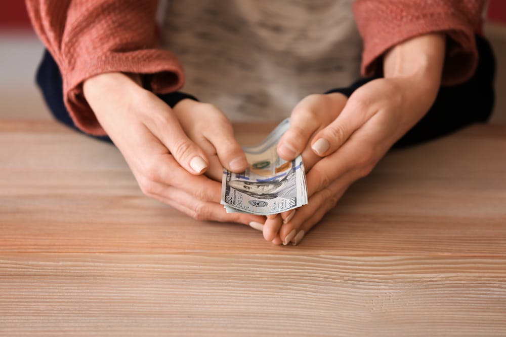 Two hands counting a stack of US dollar bills on a wooden surface, suggesting a financial transaction or money management.