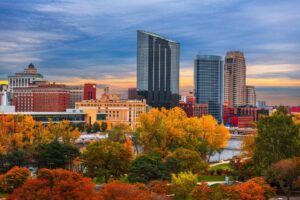 Skyscrapers tower over an urban park with autumn-colored trees, beside a calm river, under a dusky sky with soft clouds.