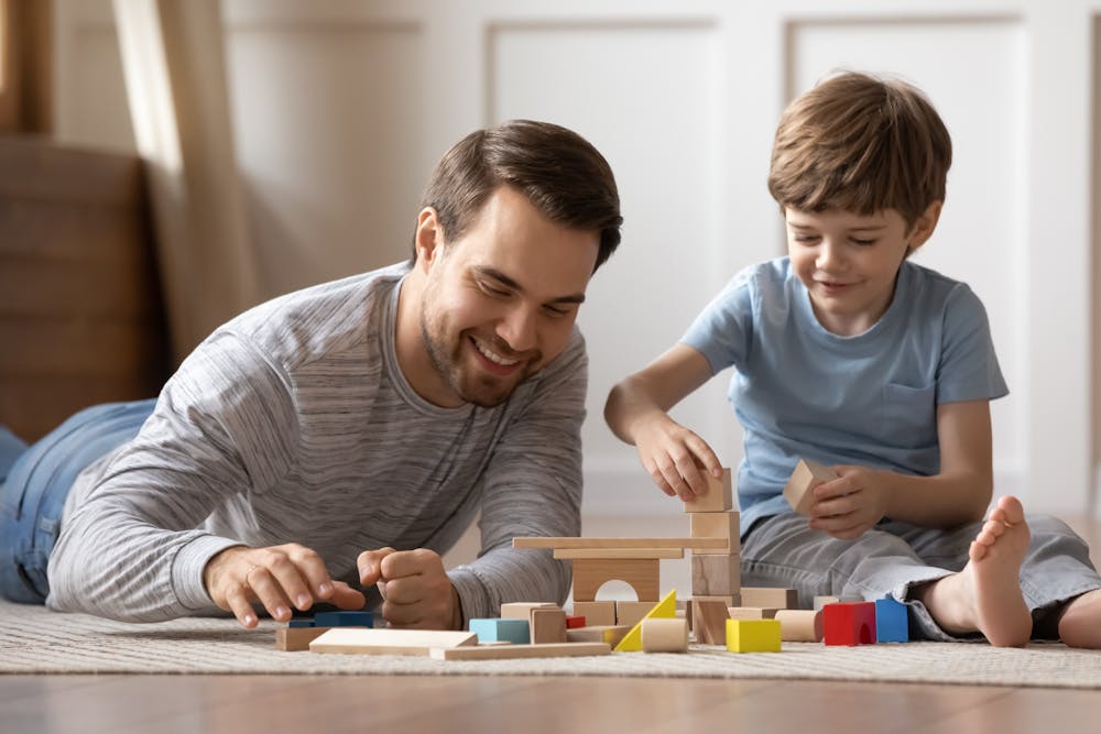 A man and a young boy are building with wooden blocks on the floor, smiling and engaged in a playful activity together indoors.