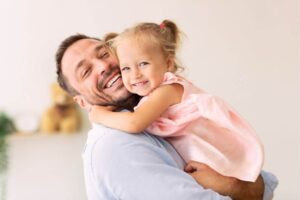 A man is hugging a young girl with affection, both smiling, in a bright room, possibly at home.