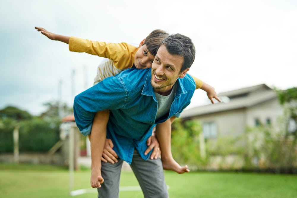 A child is piggyback riding on an adult male, both smiling, in a grassy backyard with a house in the distance.