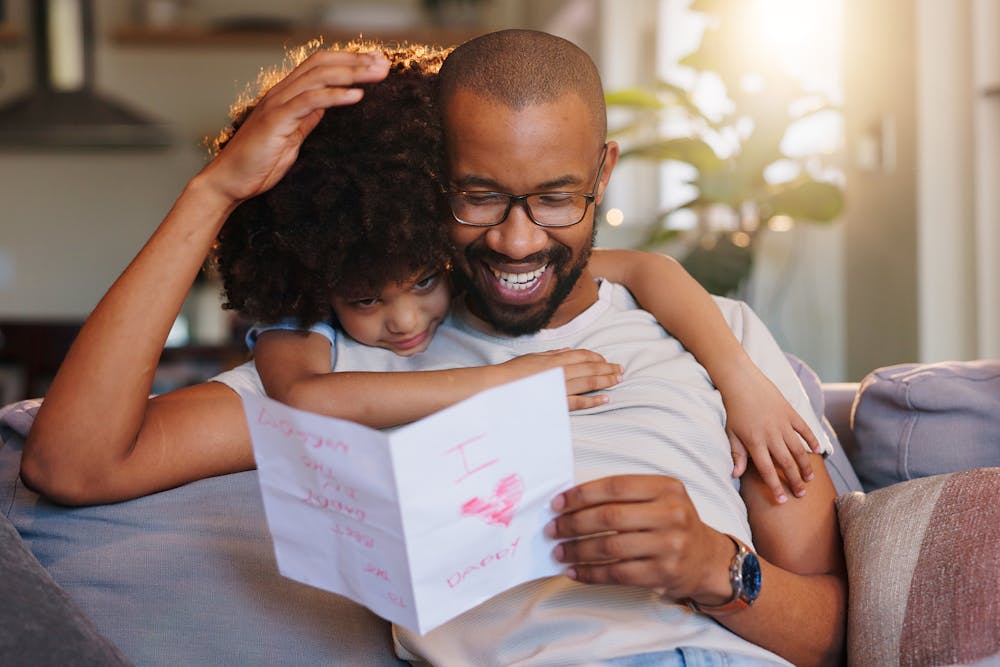 A smiling man hugs a child who holds a handmade card reading "Love you Daddy," implying affection and family bonding in a cozy indoor setting.