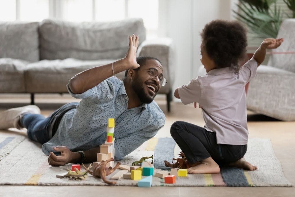 A man and child play on a rug with building blocks, simulating a high-five, in a cozy living room.