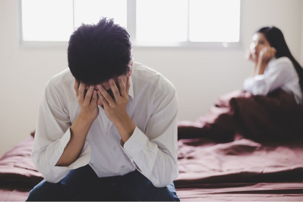 A man with his head in his hands sits at the edge of a bed, facing away from a woman in the background who is also sitting on the bed, blurred. They appear emotionally distressed in a brightly lit room.