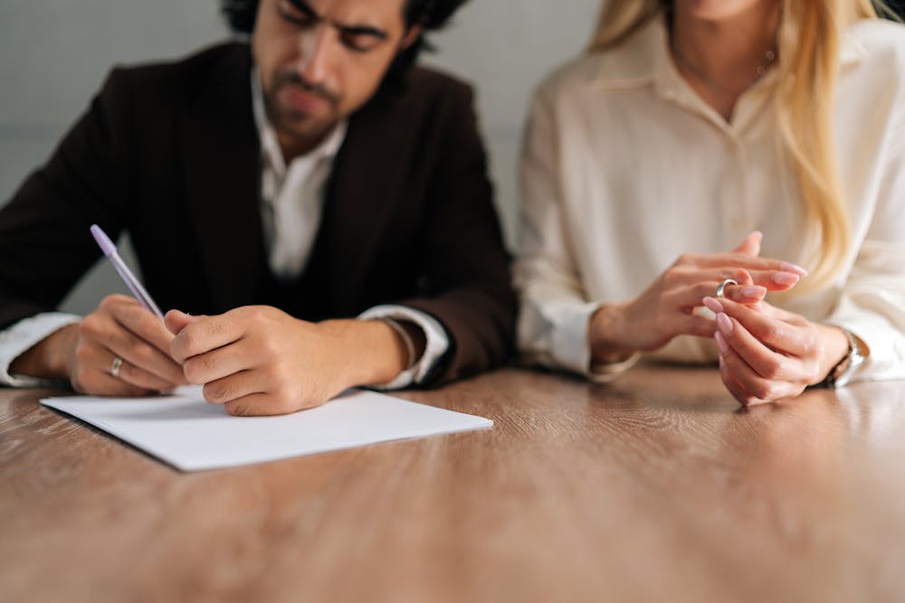 A man writes on a paper beside a woman with interlocked fingers, suggesting a business or formal setting with a wooden table foreground.