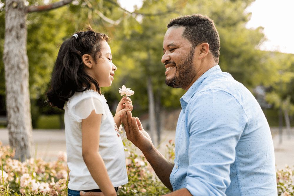 A father crouching down to his daughter's level in a sunny park