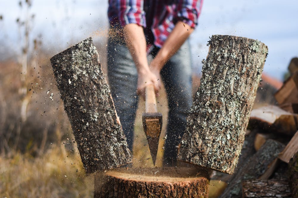 An axe splits a log mid-air on a chopping block, with wood chips scattered around, amidst a rustic outdoor setting.