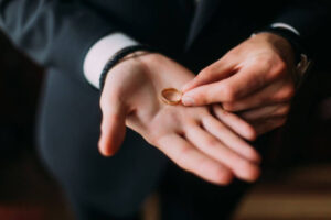 A person in a dark suit holds a golden wedding ring delicately between two fingers, likely symbolizing a moment related to a marriage ceremony.