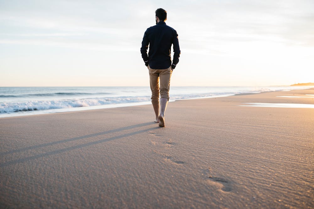 A person walks away barefoot on a sandy beach, leaving footprints, with waves and sunrise in the background.