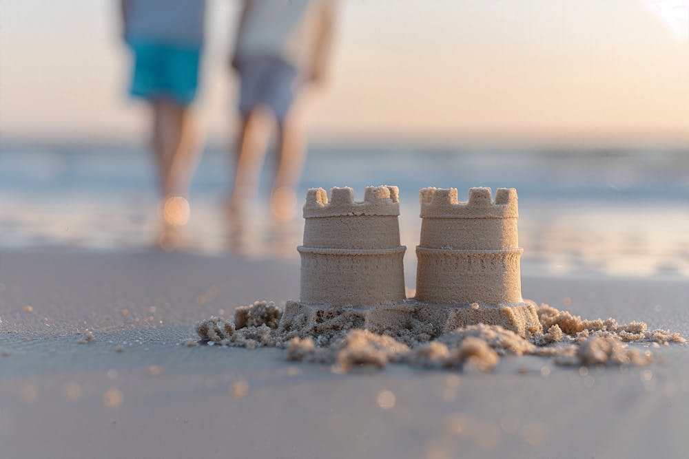 Two small sandcastles on a beach foreground, with blurred figures walking in the background during what appears to be sunrise or sunset.