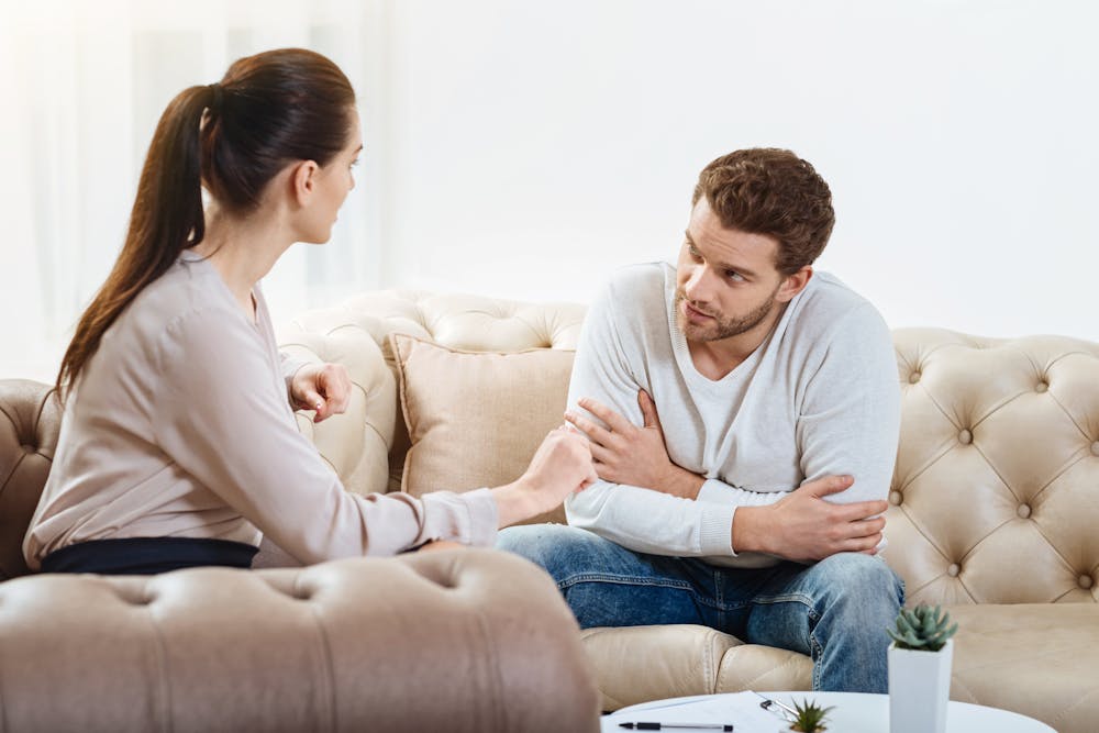 A man and woman converse earnestly on a beige sofa; the man looks concerned while the woman gestures, a notepad and pen on the table in front of them.