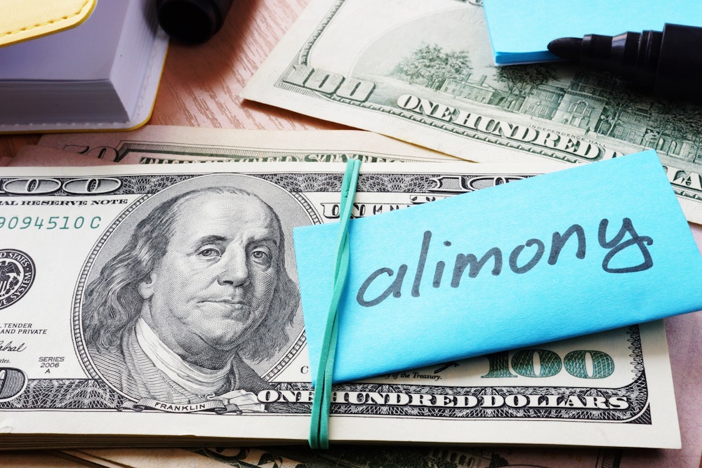 Stack of U.S. 100 dollar bills bound together with a blue rubber band labeled "alimony" on a sticky note, symbolizing financial obligations post-divorce.