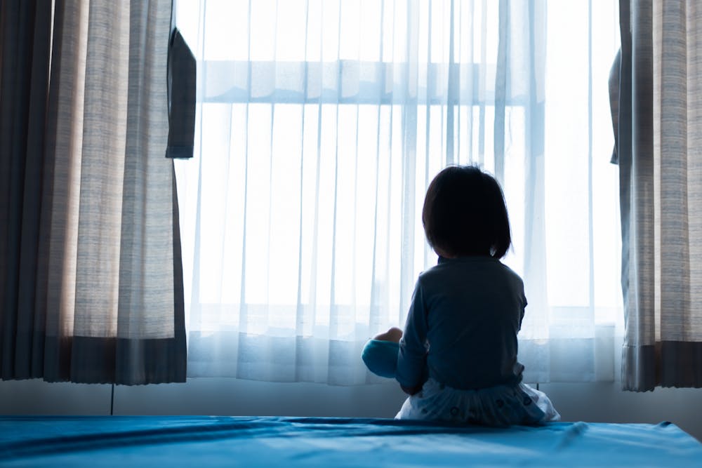 A child sits facing a sheer-curtained window, silhouetted by daylight, in an indoor setting.