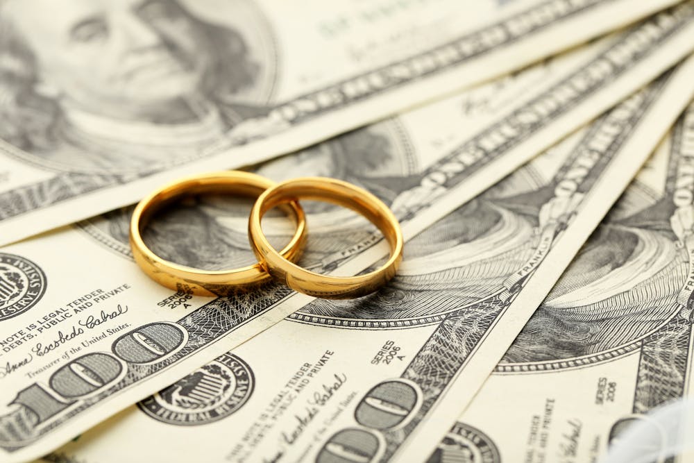 Two gold wedding rings rest on a spread of US hundred-dollar bills, symbolizing a union or marriage intertwined with wealth or finances.