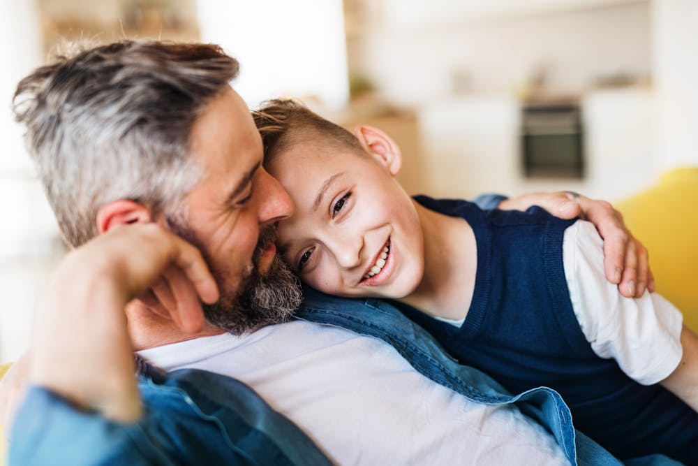 A man and a boy embrace, smiling warmly in a cozy, brightly-lit room. Their expression shows affection and happiness in a relaxed, domestic setting.